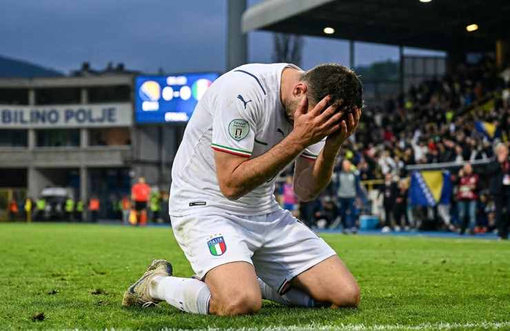 Calciatore dell'Italia in maglia bianca in ginocchio disperato allo stadio Bilino Polje dopo l'eliminazione dai Mondiali 2026.