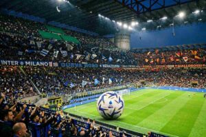 Rapresentazione realistica dello stadio Giuseppe Meazza con tifosi dell'Inter e della Roma, il pallone della Serie A al centro della scena e parte del campo.